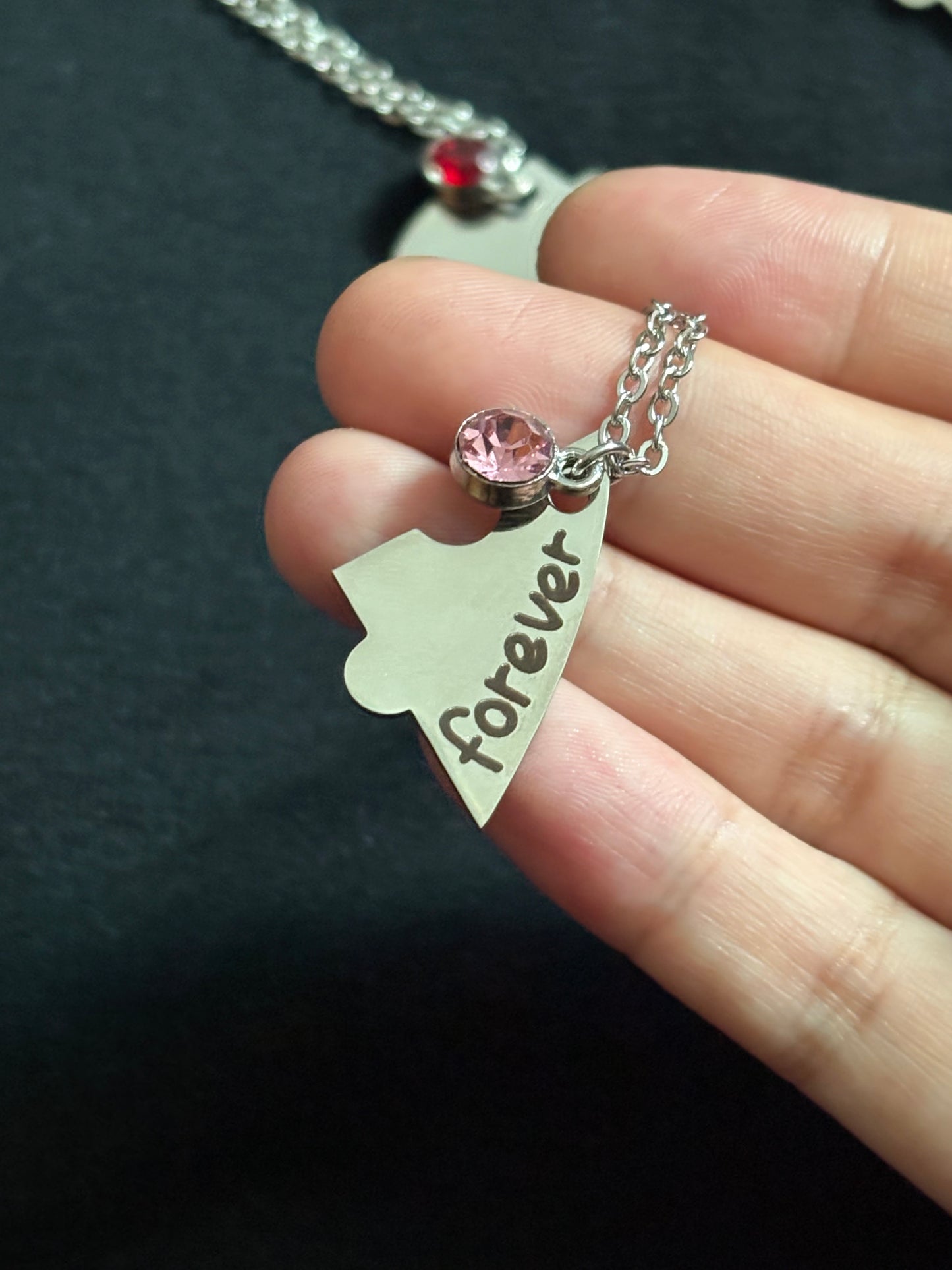 Heart-shaped pendant with 'forever' engraving and pink gemstone held between fingers against a dark background