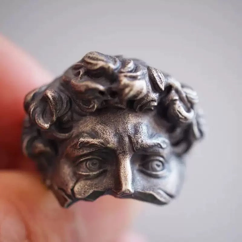 Bronze-colored ring with intricate design held between fingers against a neutral background